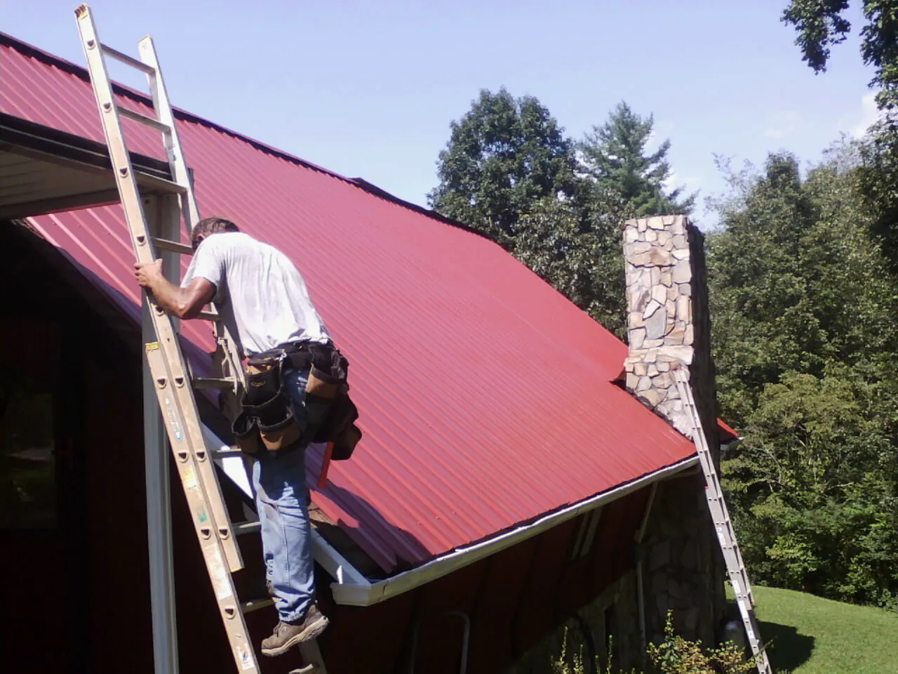 Roofer on a ladder beside a red metal roof and stone chimney — Soffit & Fascia Repair by Roofing Brothers in New Freedom